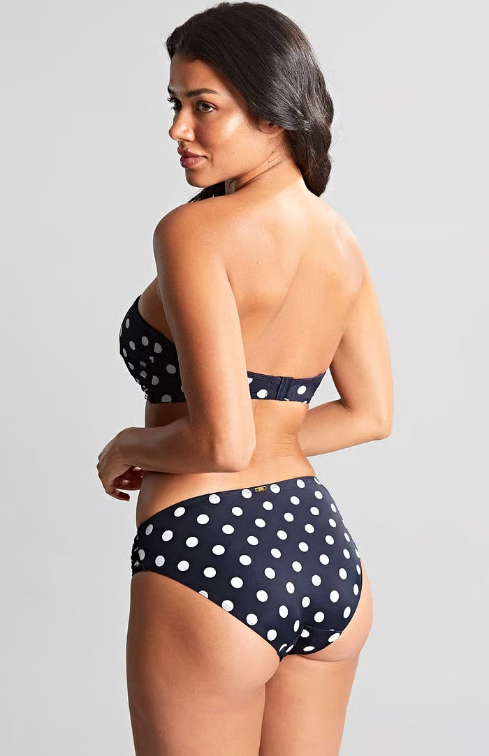 A woman modeling a two-piece bikini set with a navy blue and white polka dot pattern, shown from a three-quarters back view against a light gray studio background.