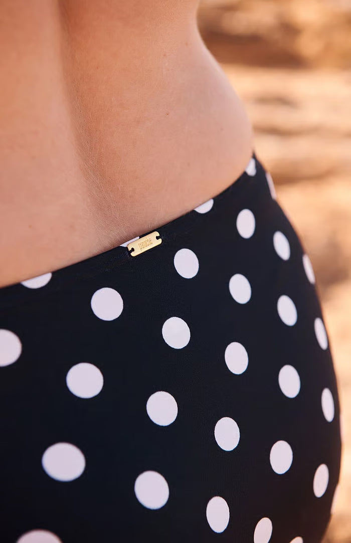 Close-up of a person wearing black swim bottoms with white polka dots and a small gold brand tag at the waistband, against a blurred sandy background.
